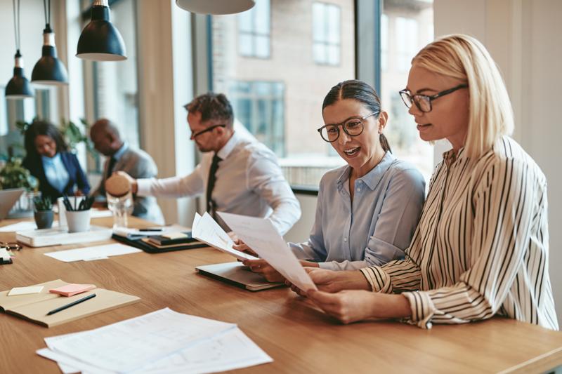 Two smiling young businesswomen reading paperwork together while sitting at a boardroom table during an office meeting