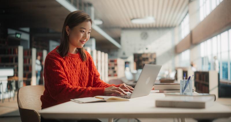 Engaged Asian Female Student Working on Her University Assignments With a Laptop in Modern Library. Young Woman in Red Sweater Studying, Surrounded by Books and Pens.