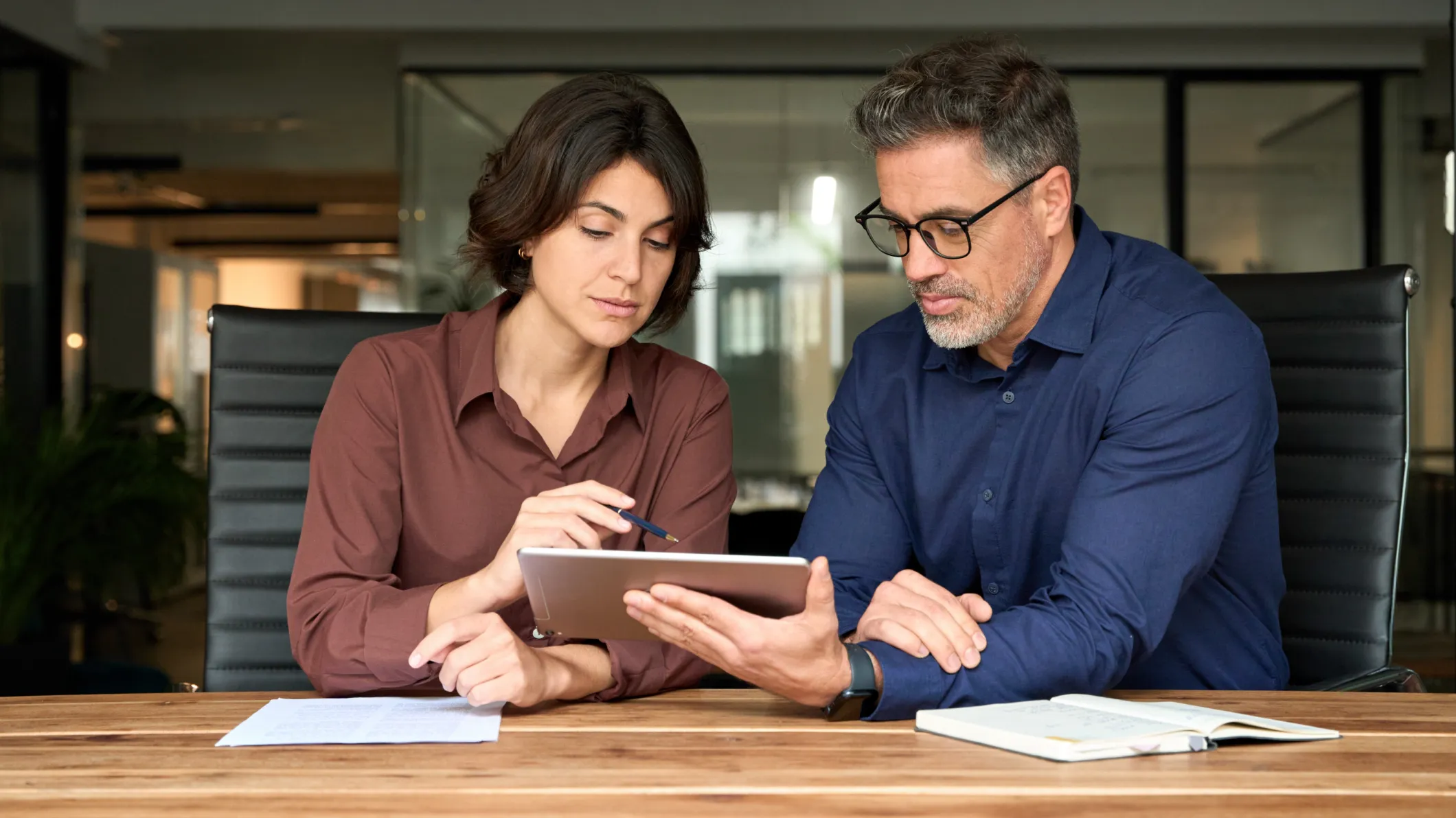 A woman and a man sitting at a desk looking at a tablet.
