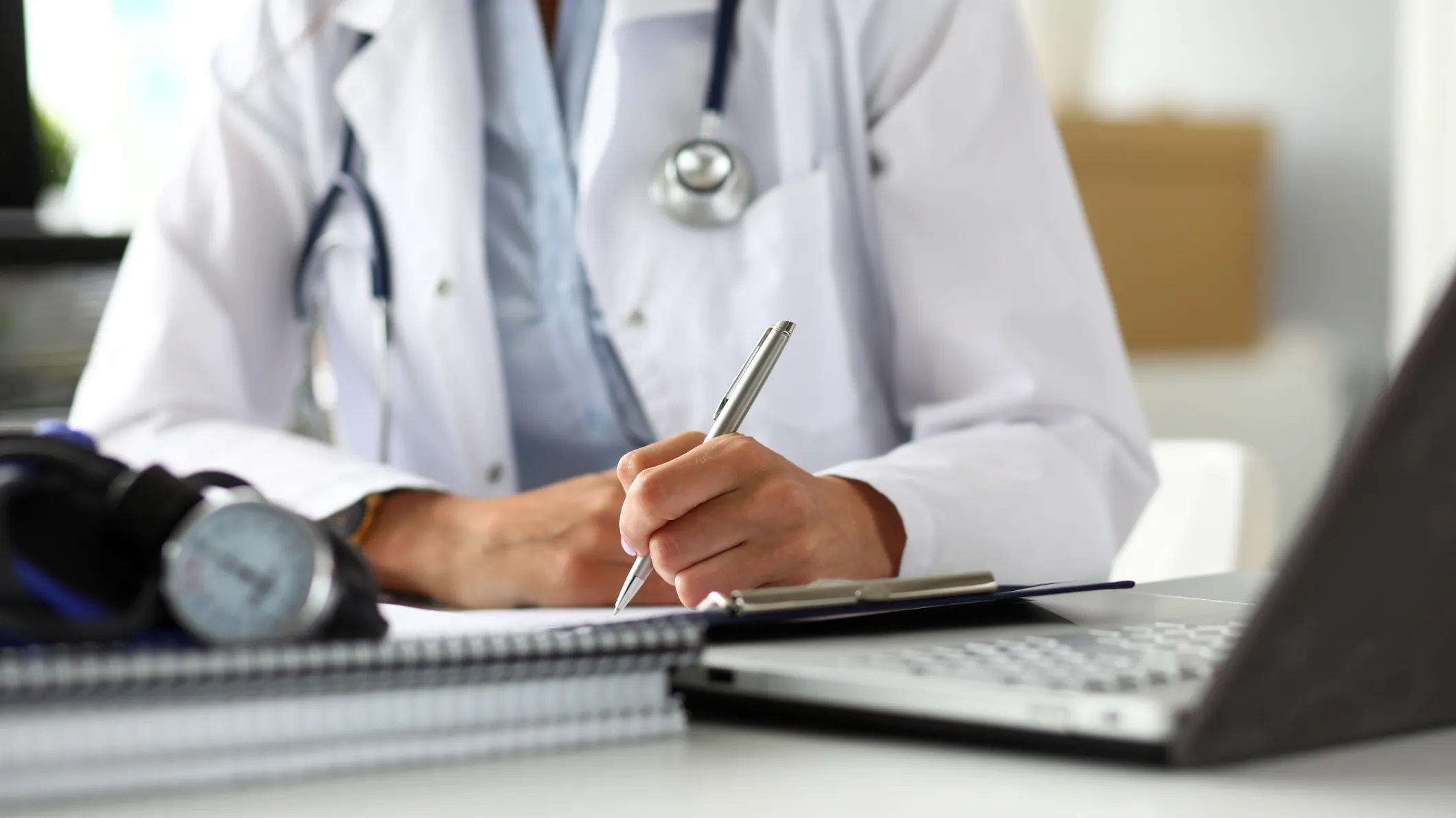 A doctor writes on a clipboard while sitting at a desk with an open laptop