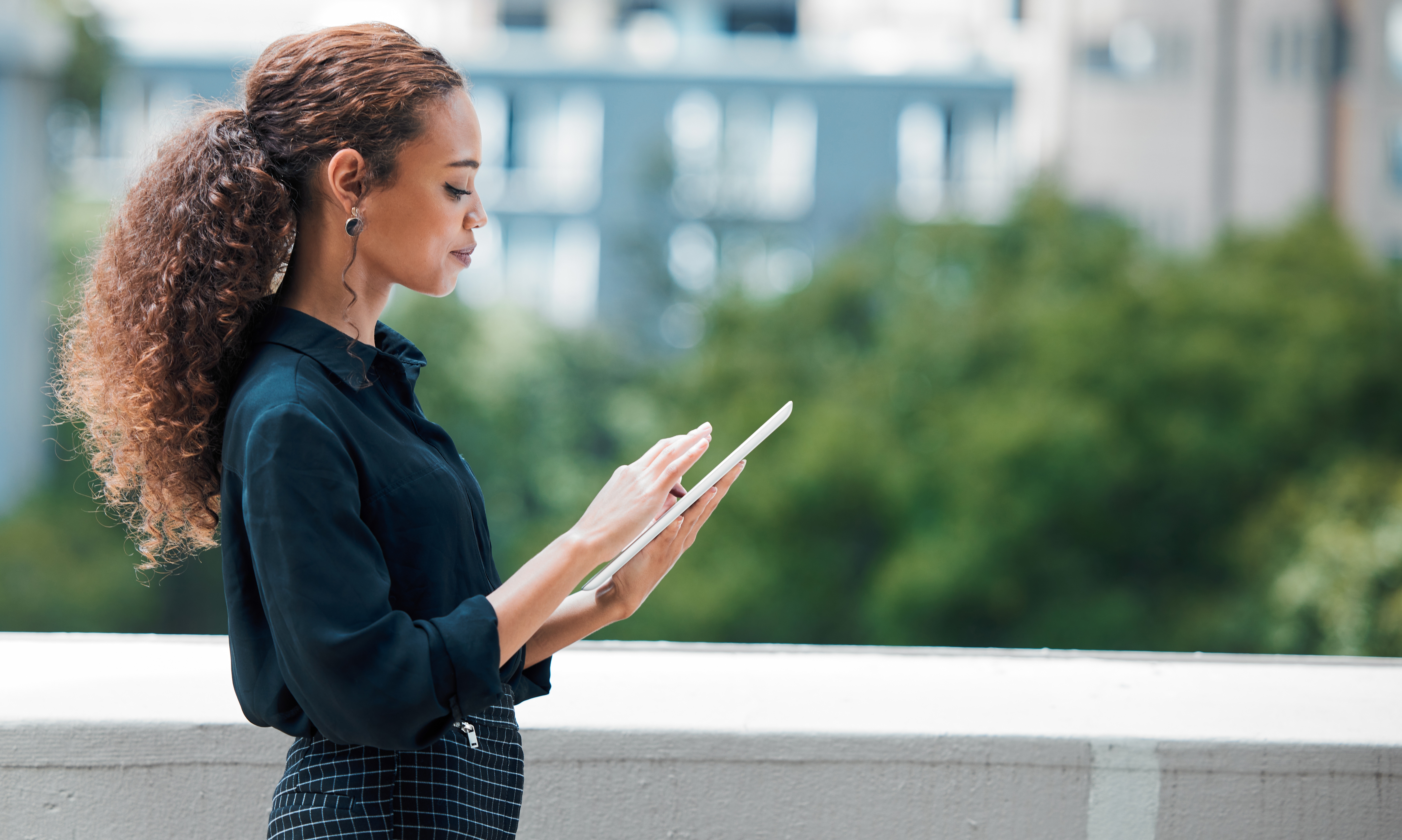 A woman with long, curly hair (which is tied back) checks something on a tablet computer. She is facing to the viewer's right.