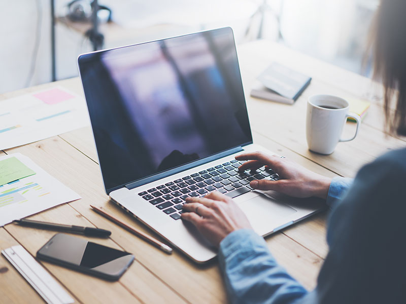 Woman sat at desk using Financial Management software for accounting on laptop with coffee, pens, note book and phone surrounding her